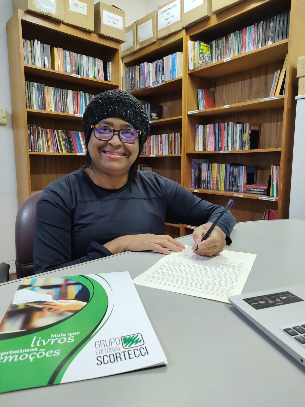 Cida Agnes assinando contrato na biblioteca, com material do Grupo Editorial Scortecci